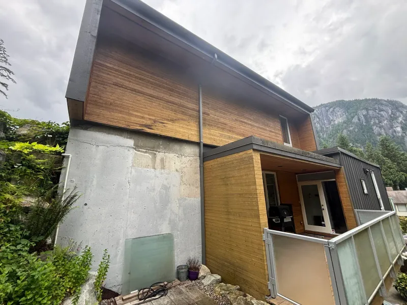 Before: Side elevation of house showing siding above a concrete foundation wall, with windows and a covered deck below, . Arbour woods, Squamish, BC