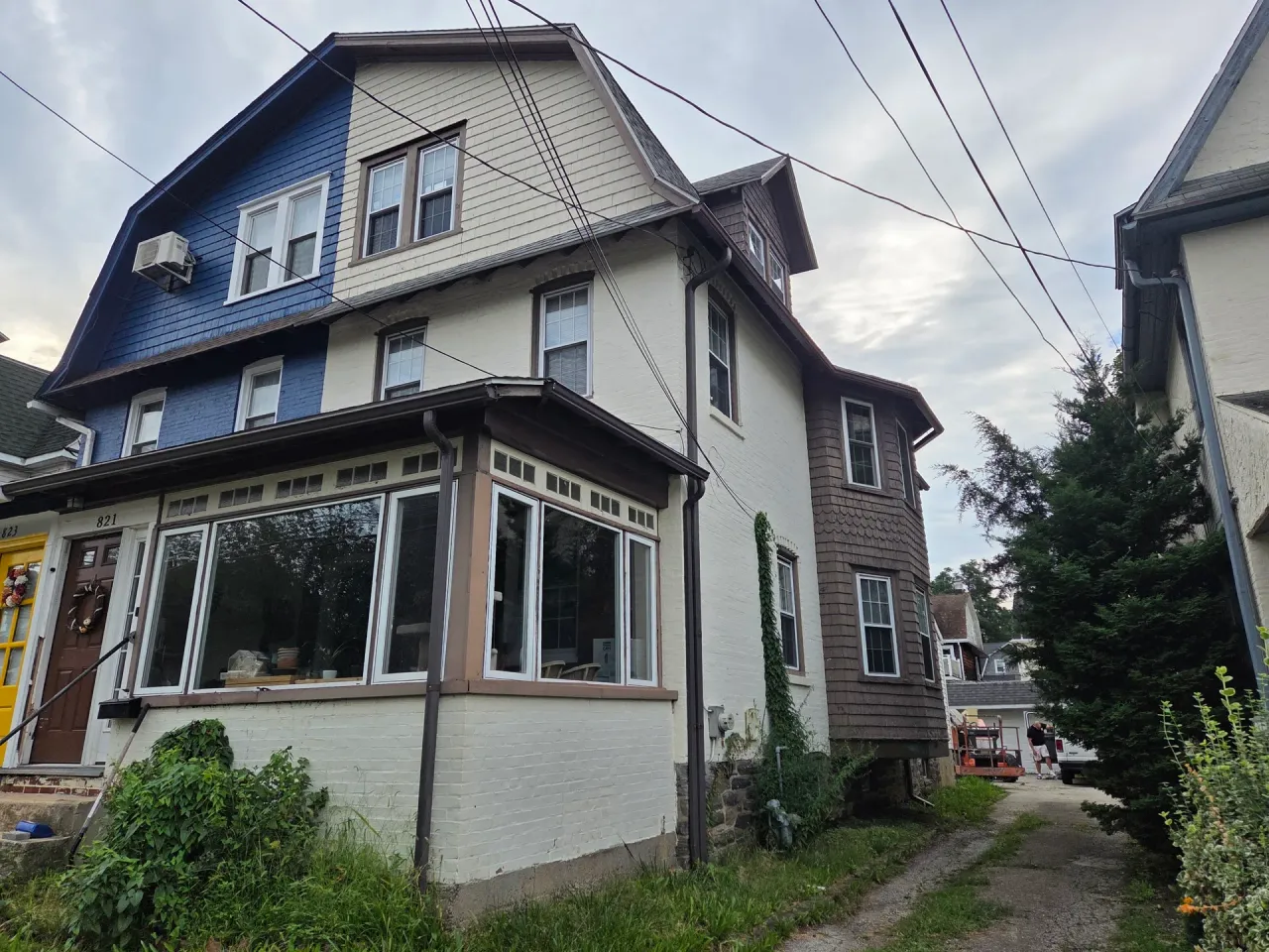 Before: Front corner view of a two-and-a-half story house with a sunroom with large windows, a bay window tower, and a dormered upper roof, before painting., Ardmore Lower Merion, PA