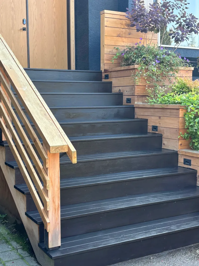 After: Close angle of front staircase with dark painted treads and risers, handrail, and planter boxes with flowering plants, Highlands, Squamish, BC