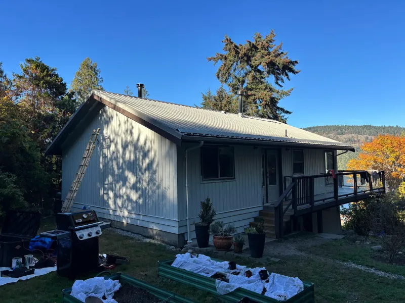 After: Rear elevation of house with freshly painted cream siding, metal roof, deck, and railing, Souther Salt Spring. Isabella Point / Roland Rd, BC