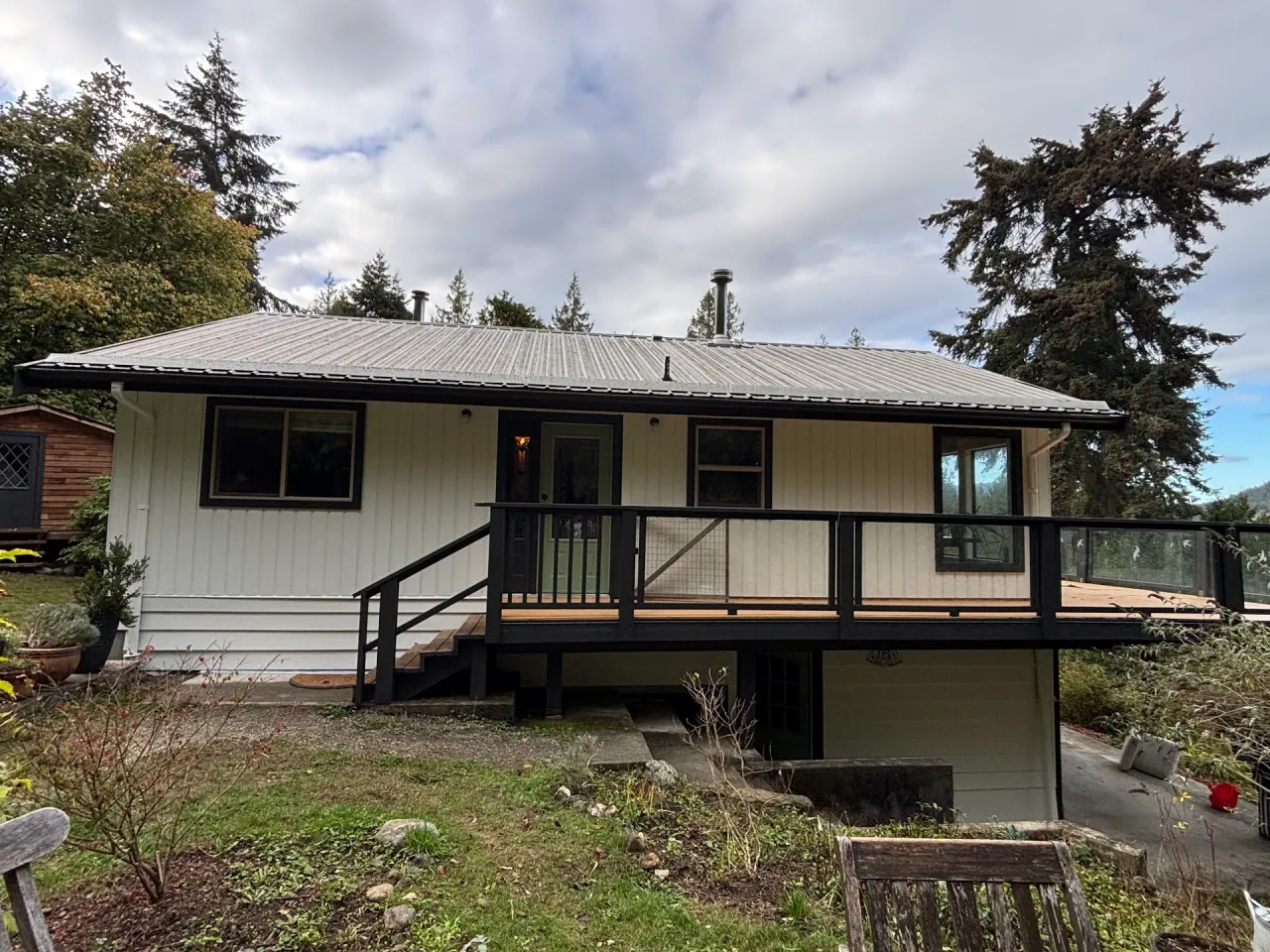 After: Rear elevation of house with cream vertical siding, dark trim, metal roof, and deck with railing, Souther Salt Spring. Isabella Point / Roland Rd, BC