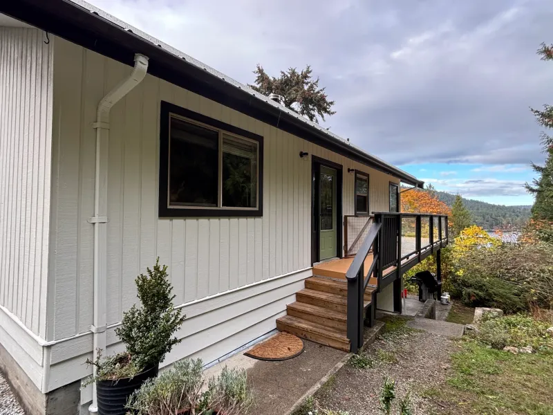 After: Rear elevation of house with cream siding, dark trim, door, window, and deck with stairs, Souther Salt Spring. Isabella Point / Roland Rd, BC