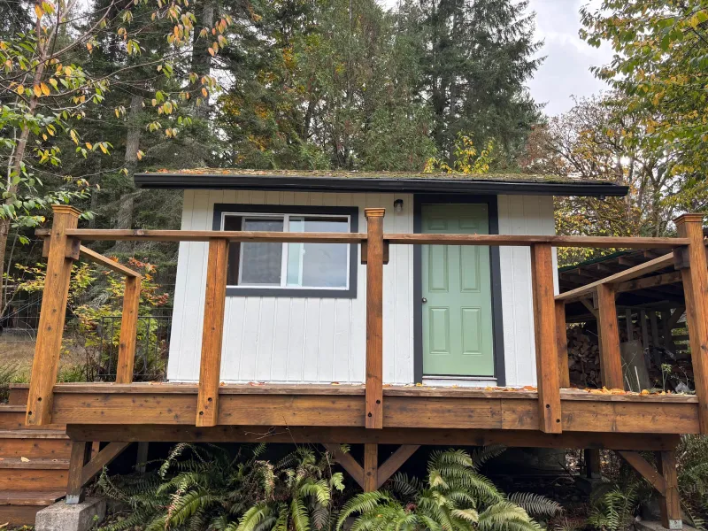 After: Rear elevation of small cabin with white siding, dark trim, green door, and deck railing, Souther Salt Spring. Isabella Point / Roland Rd, BC