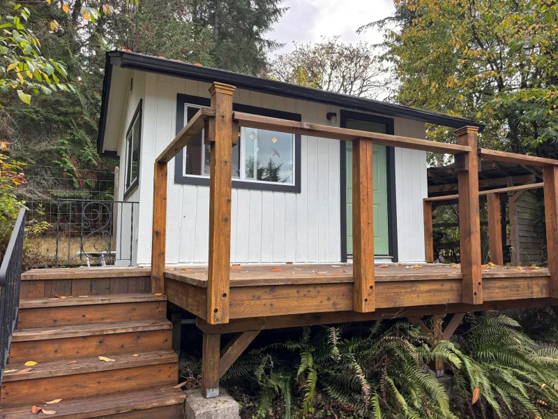 After: Side view of small cabin with white siding, dark trim, green door, and deck, Souther Salt Spring. Isabella Point / Roland Rd, BC