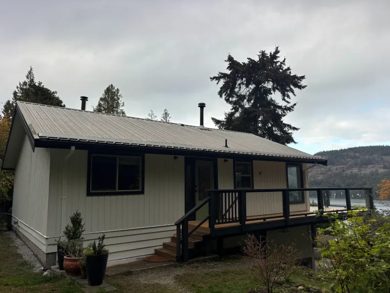 After: Front elevation of house with cream vertical siding, dark trim, metal roof, deck, and lake view beyond, Souther Salt Spring. Isabella Point / Roland Rd, BC