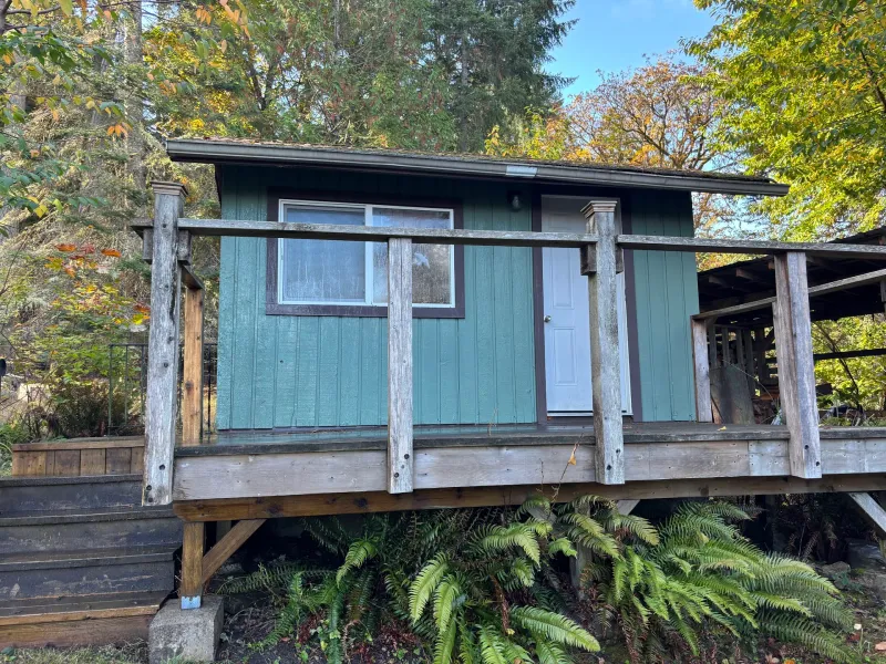 Before: Rear elevation of small teal cabin with white door, window, and deck railing, Souther Salt Spring. Isabella Point / Roland Rd, BC