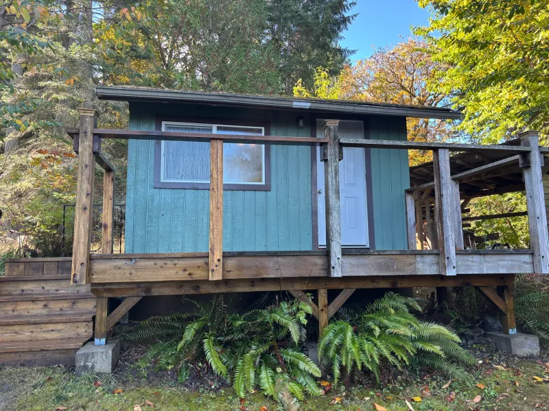 Before: Rear elevation of teal cabin with window, white door, and deck railing, Souther Salt Spring. Isabella Point / Roland Rd, BC